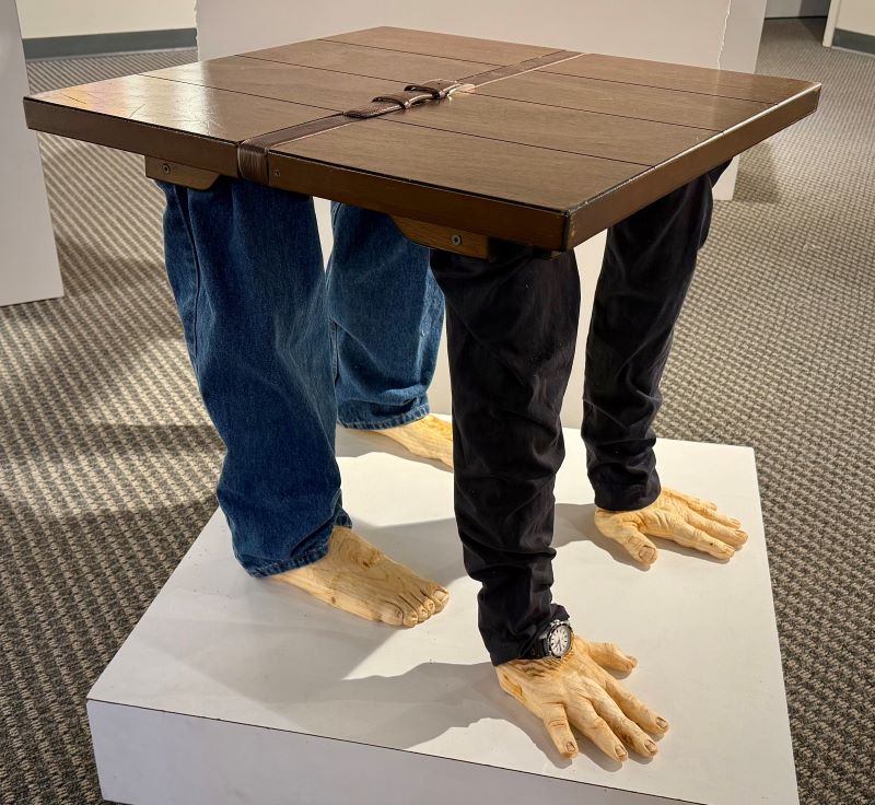 A table with human arms and human legs shaped in wood as table legs. The table wears sleeves and jeans.