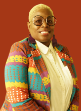 A photo of Nola D. Oracle. She is a Black woman with short hair. She wears large, circular glasses and a colorful blazer.