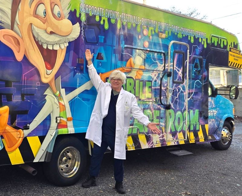 Man dressed as a mad scientist stands outside the Escape Me Mobile bus, which also features a cartoon scientist on the vehicle.