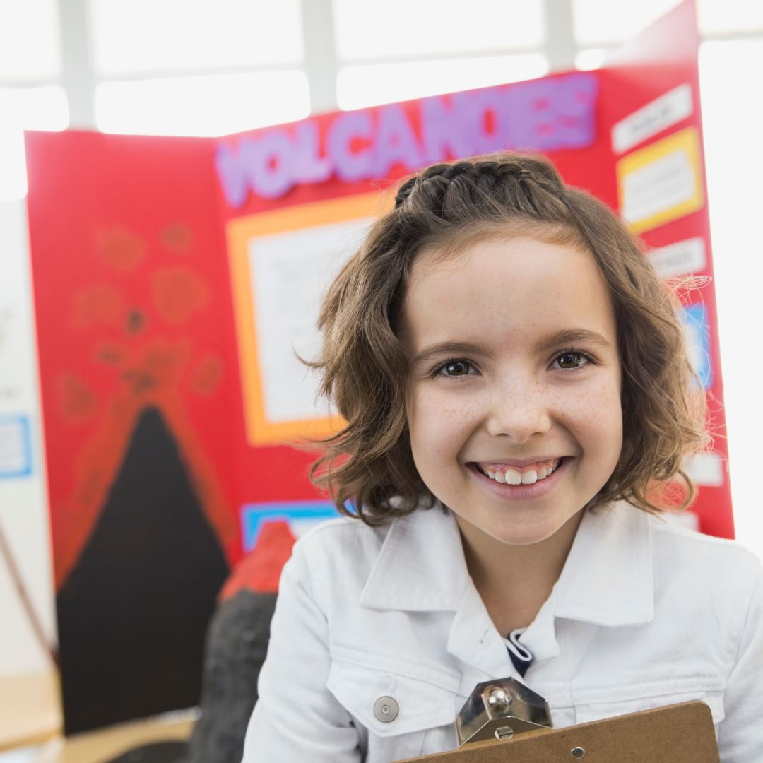 young girl smiles in front of a science fair display on volcanoes
